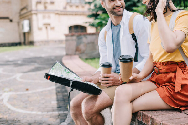 cropped view of happy bearded man with paper cup and map sitting near girl 