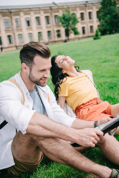 selective focus of cheerful man holding map near woman sitting on grass 