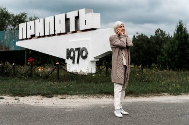 PRIPYAT, UKRAINE - AUGUST 15, 2019: retired woman standing near monument with pripyat letters and covering ears  clipart