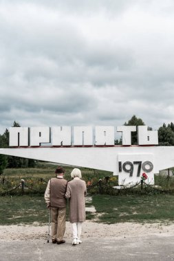 PRIPYAT, UKRAINE - AUGUST 15, 2019: back view of retired husband and wife standing near monument with pripyat letters clipart
