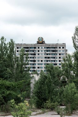 PRIPYAT, UKRAINE - AUGUST 15, 2019: green trees near building against sky with clouds in chernobyl  clipart