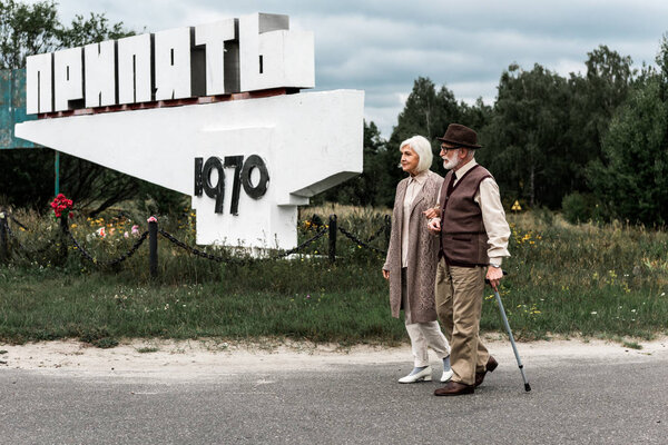 PRIPYAT, UKRAINE - AUGUST 15, 2019: retired man and woman walking near monument with pripyat letters