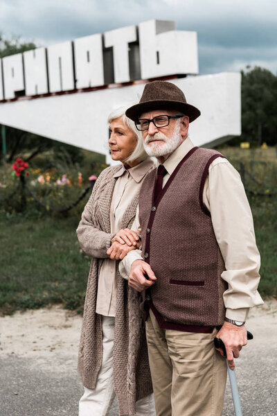 PRIPYAT, UKRAINE - AUGUST 15, 2019: retired husband and wife standing with walking cane near monument with pripyat letters
