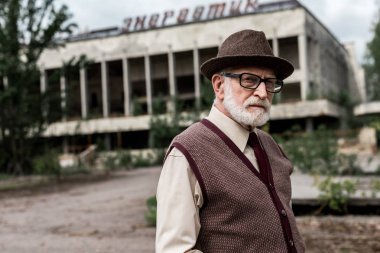 Pripyat, Ukrayna - 15 Ağustos 2019: Çernobil'de enerjik harflerle binanın yakınında bagaj tutan gözlüklü kıdemli adam 