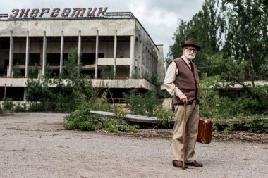 Pripyat, Ukrayna - 15 Ağustos 2019: Çernobil'de enerjik harflerle binanın yakınında bagaj tutan şapkalı kıdemli adam 