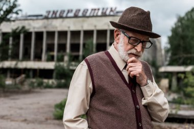 PRIPYAT, UKRAINE - AUGUST 15, 2019: pensive senior man standing near building with energetic lettering in chernobyl  clipart