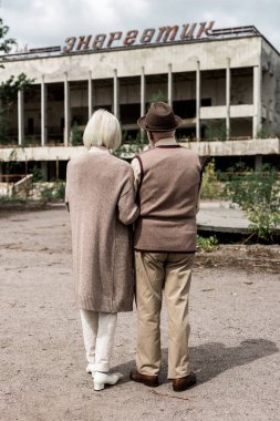 PRIPYAT, UKRAINE - AUGUST 15, 2019: back view of retired couple standing near building with energetic lettering in chernobyl  clipart