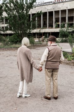 PRIPYAT, UKRAINE - AUGUST 15, 2019: back view of retired man and woman holding hands near building  clipart