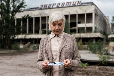 PRIPYAT, UKRAINE - AUGUST 15, 2019: senior woman looking at photo near building with energetic lettering in chernobyl  clipart