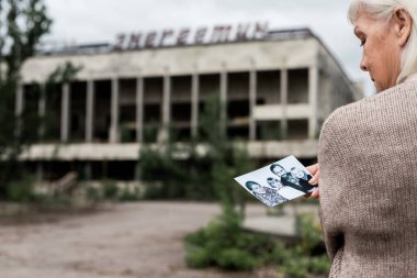 PRIPYAT, UKRAINE - AUGUST 15, 2019: selective focus of woman with grey hair holding photo near building with lettering in chernobyl  clipart