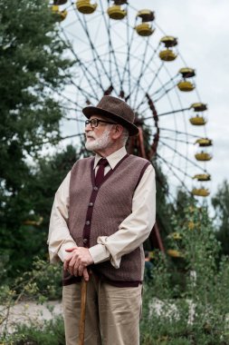 PRIPYAT, UKRAINE - AUGUST 15, 2019: bearded senior man in hat standing with walking cane in amusement park with ferris wheel 