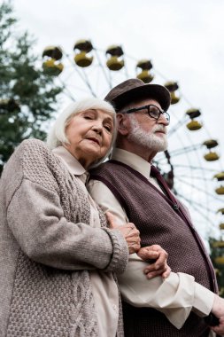 Pripyat, Ukrayna - 15 Ağustos 2019: gözlüklü sakallı yaşlı adamın eğlence parkında dönme dolabın yanında eşiyle ayakta durması
