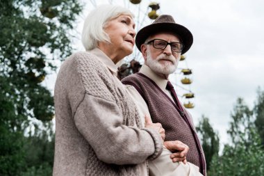 Pripyat, Ukrayna - 15 Ağustos 2019: Lunaparkta dönme dolap yakınında emekli karısıyla ayakta duran sakallı yaşlı adamın düşük açı görünümü