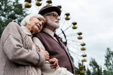 Pripyat, Ukrayna - 15 Ağustos 2019: Lunaparkta dönme dolap yakınında kocasına sarılan yaşlı kadının düşük açılı görünümü