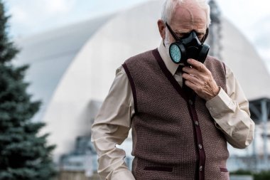 Pripyat, Ukrayna - 15 Ağustos 2019: koruyucu maskeye dokunan ve terk edilmiş çernobil reaktörünyakınında duran kıdemli adam 