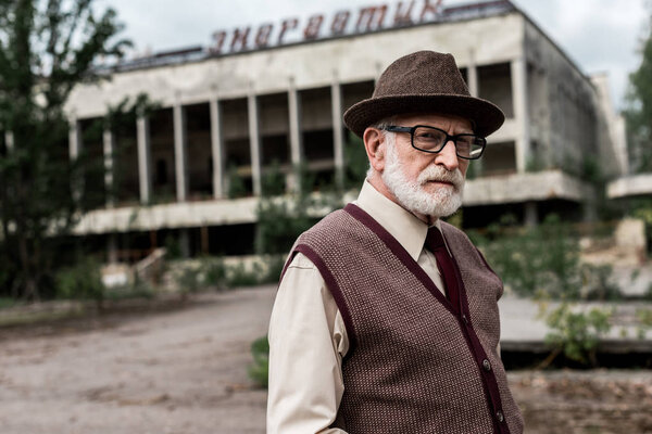 PRIPYAT, UKRAINE - AUGUST 15, 2019: senior man in glasses holding luggage near building with energetic lettering in chernobyl 
