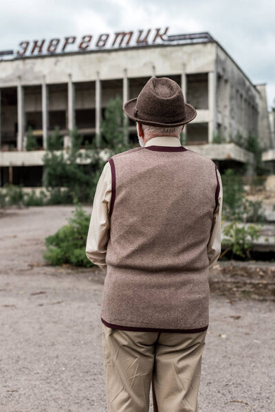 PRIPYAT, UKRAINE - AUGUST 15, 2019: back view of senior man in hat standing near building with energetic lettering in chernobyl 