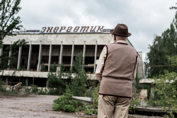 PRIPYAT, UKRAINE - AUGUST 15, 2019: back view of retired man in hat standing near building with energetic lettering in chernobyl 