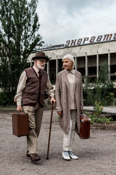 PRIPYAT, UKRAINE - AUGUST 15, 2019: retired couple with suitcases near building with energetic lettering in chernobyl 