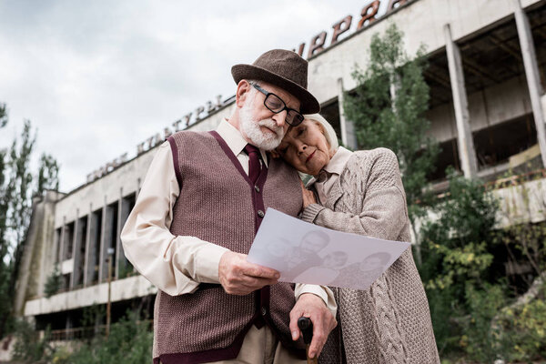 ПРИПЯТ, УКРАИНА - 15 АВГУСТА 2019: вид под низким углом пожилой пары, смотрящей на фото возле здания в Чернобыле
 