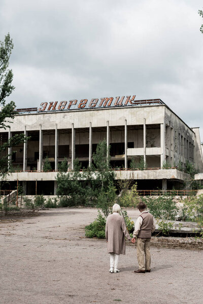 PRIPYAT, UKRAINE - AUGUST 15, 2019: back view of retired couple holding hands near building with energetic lettering in chernobyl 