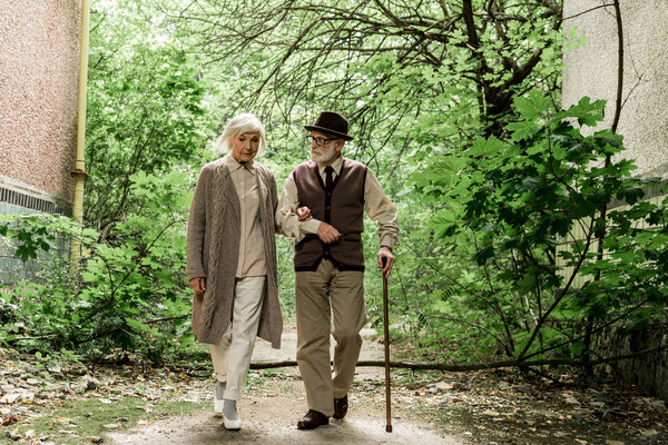 retired woman and senior man with walking cane walking near green trees 