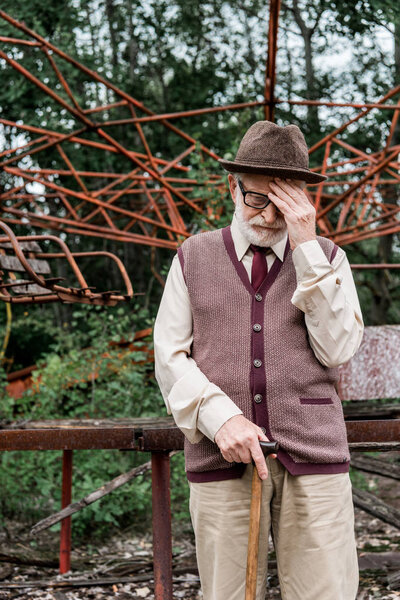 PRIPYAT, UKRAINE - AUGUST 15, 2019: bearded retired man in hat and glasses standing with walking cane near damaged carousel 