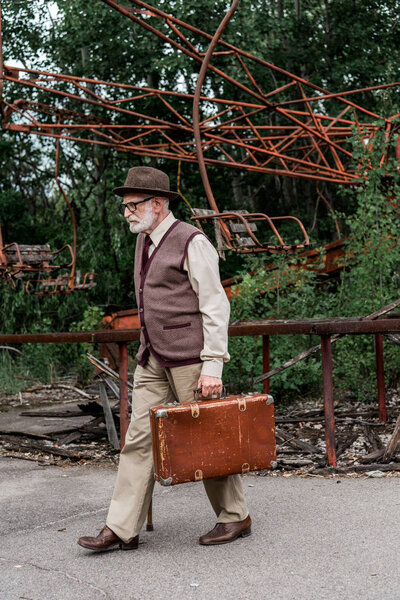 ПРИПЯТ, УКРАИНА - 15 августа 2019 года: бородатый пенсионер в шляпе и очках ходит с тростью и чемоданом рядом с поврежденной каруселью
 