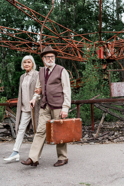 PRIPYAT, UKRAINE - AUGUST 15, 2019: senior man in glasses holding suitcase while walking with wife near abandoned carousel 