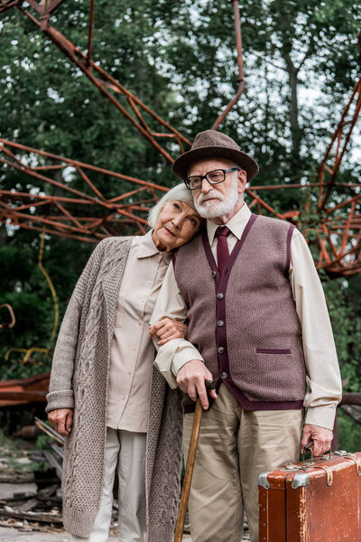 PRIPYAT, UKRAINE - AUGUST 15, 2019: senior man in glasses holding suitcase while standing with wife near abandoned carousel 