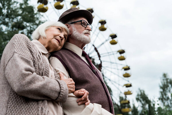 PRIPYAT, UKRAINE - AUGUST 15, 2019: low angle view of senior woman hugging husband near ferris wheel in amusement park