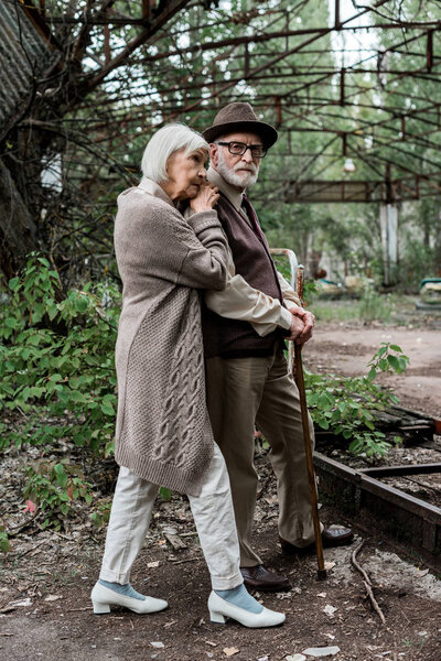 sad senior woman standing with retired husband in hat and glasses 