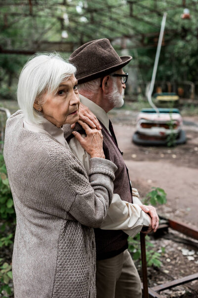 PRIPYAT, UKRAINE - AUGUST 15, 2019: senior man in hat standing with retired wife near amusement park in chernobyl 