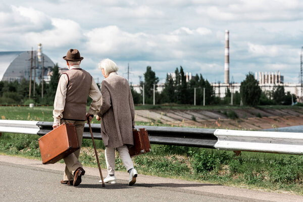 PRIPYAT, UKRAINE - AUGUST 15, 2019: back view of senior man and woman walking with baggage near chernobyl nuclear power plant 