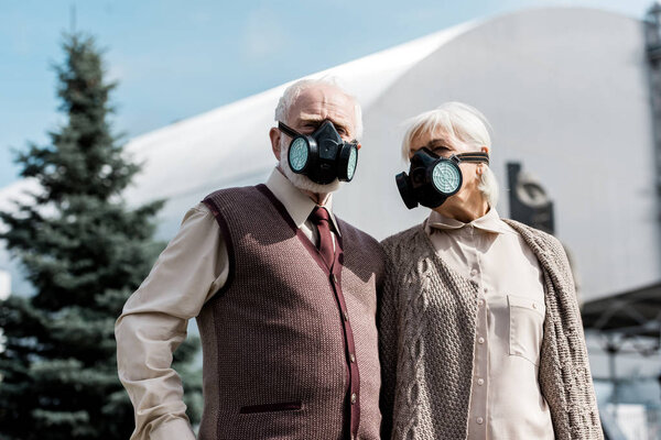 ПРИПЯТ, УКРАИНА - 15 августа 2019 года: пара пенсионеров в защитных масках стоит возле заброшенного Чернобыльского реактора
 
