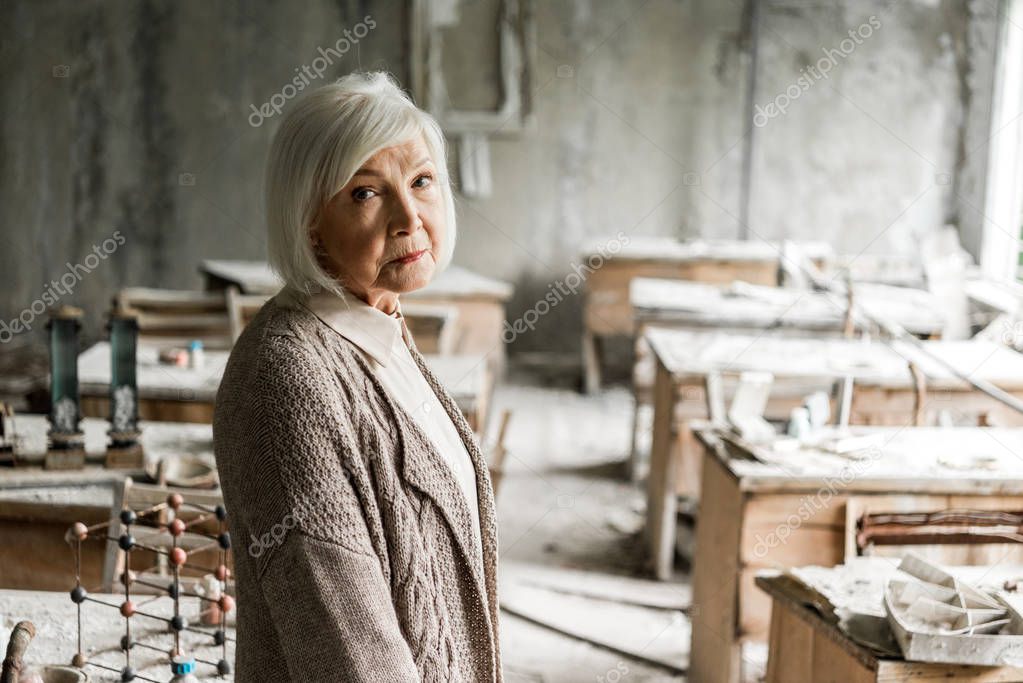 selective focus of frustrated senior woman standing in dirty abandoned classroom 