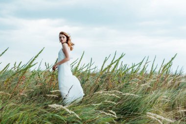 selective focus of attractive redhead woman in white dress looking at camera  clipart