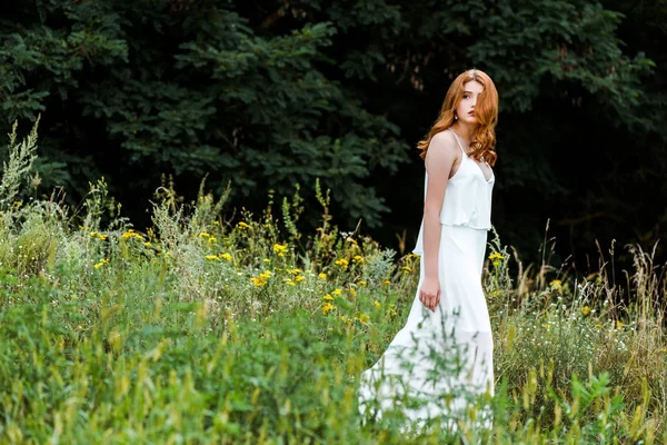 野生の花で野原を歩く白いドレスの美しい赤毛の女の子の選択的な焦点 — ストック写真