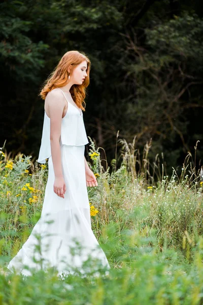 野生の花とフィールドに立っている白いドレスの赤毛の女の子の選択的な焦点 — ストック写真