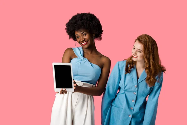 快乐红发女孩看着非洲美国妇女拿着数字平板电脑与空白屏幕隔离在粉红色 — 图库照片
