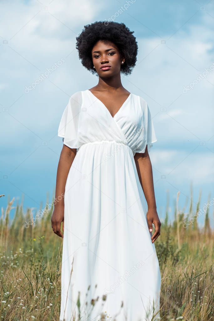 attractive and curly african american woman in white dress against blue sky 
