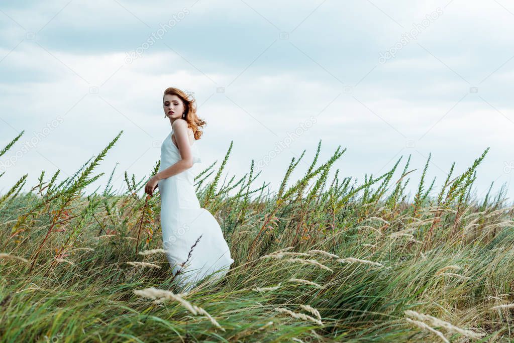 selective focus of attractive redhead woman in white dress looking at camera 