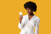 shocked curly african american woman looking at smartphone isolated on orange 