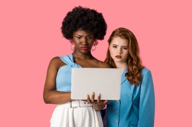 displeased african american woman looking at laptop with redhead girl isolated on pink  clipart