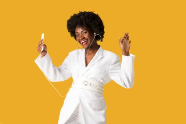 gülümseyen afro-amerikan kız akıllı telefon tutarken müzik dinlerken ve turuncu izole gesturing 