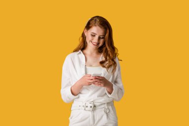 happy redhead girl using smartphone and smiling isolated on orange 