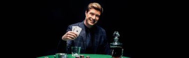 KYIV, UKRAINE - AUGUST 20, 2019: panoramic shot of happy man holding playing cards near alcohol drink isolated on black  clipart
