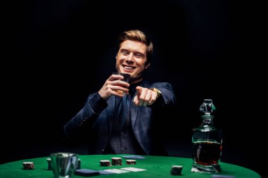 selective focus of happy man pointing with finger while holding glass with whiskey isolated on black  clipart