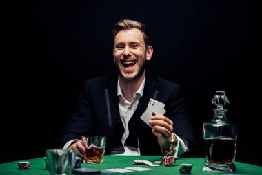 KYIV, UKRAINE - AUGUST 20, 2019: selective focus of cheerful man holding glass with alcohol and playing cards isolated on black  clipart