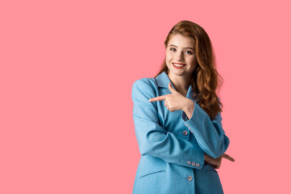 smiling redhead woman pointing with finger isolated on pink 